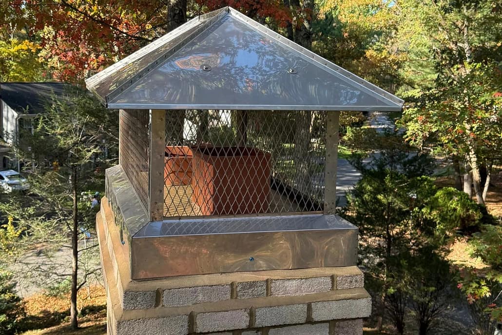 2_Stainless_Stell_Full_Coverage_CHimney_Cap_Install_Reston_VA_-__Sixpenny_Chimney_Sweeps Metal chimney cap with diamond mesh guards atop brick chimney, surrounded by autumn trees.