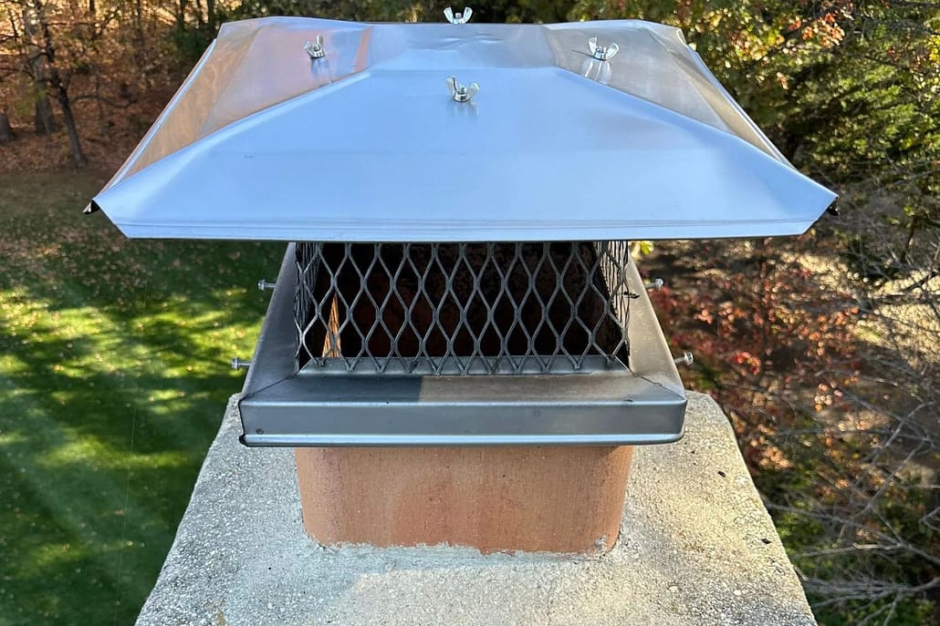 New_Flue_Chimney_Cap_Installation_Great_Falls_VA_-_Sixpenny_Chimney_Sweeps Metal chimney vent with diamond mesh guard and tilted metal cap atop brick chimney, surrounded by trees.