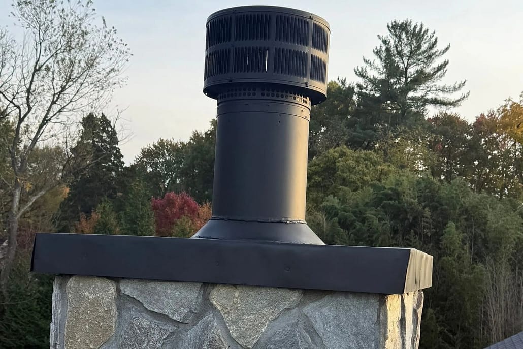 Pre-Fabricated_Chimney_Cap_Installation_Mclean_VA Chimney vent with cylindrical cap atop stone chimney, metal shielding, and forested autumn backdrop.