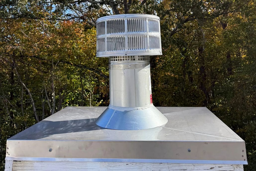 Pre-fabricated_chimney_chase_cover_and_spark_arrestor_install_Woodbridge_VA_-__Sixpenny_Chimney_Sweeps Stainless metal vent chimney rises from flat metal roof, surrounded by autumn trees in background.