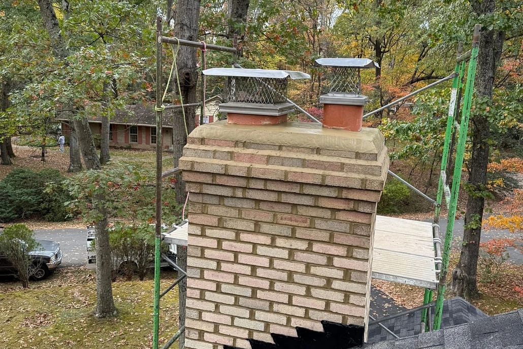 Scaffolding_against_chimney_during_repairs_in_reston_va_-__Sixpenny_Chimney_Sweeps Brick chimney with two metal caps and mesh guards, surrounded by autumn trees and scaffold.
