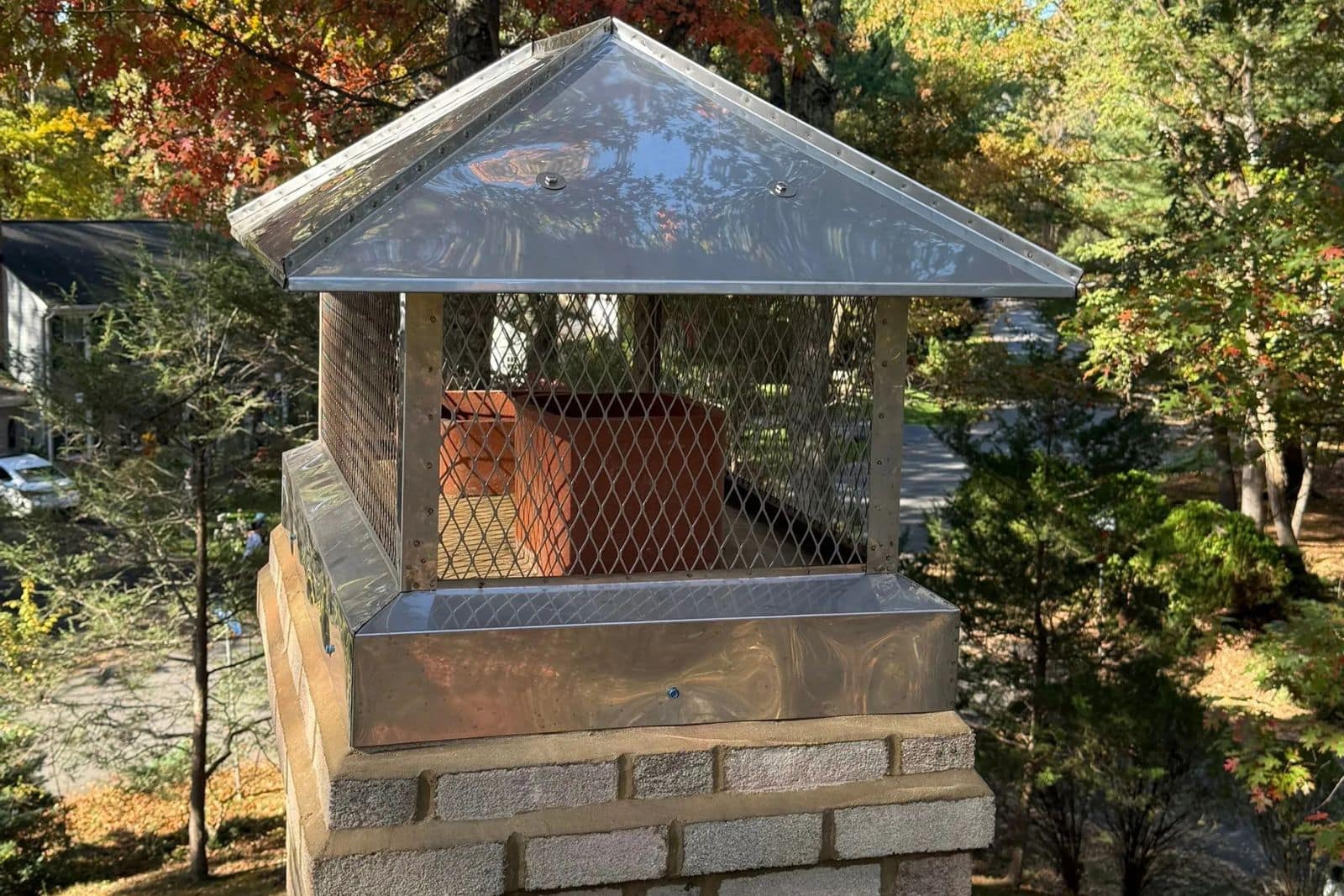2_Stainless_Stell_Full_Coverage_CHimney_Cap_Install_Reston_VA_-__Sixpenny_Chimney_Sweeps Metal chimney cap with diamond mesh guards atop brick chimney, surrounded by autumn trees.