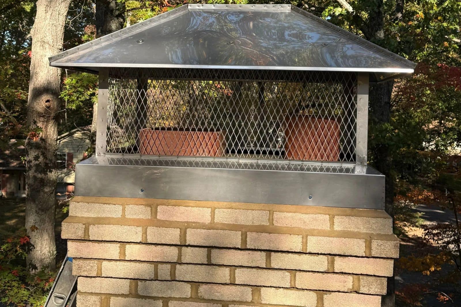 Completed_Chimney_Repair__New_Full_coverage_cap_reston_va Metal chimney cap with mesh guard atop brick chimney, surrounded by trees and autumn foliage.
