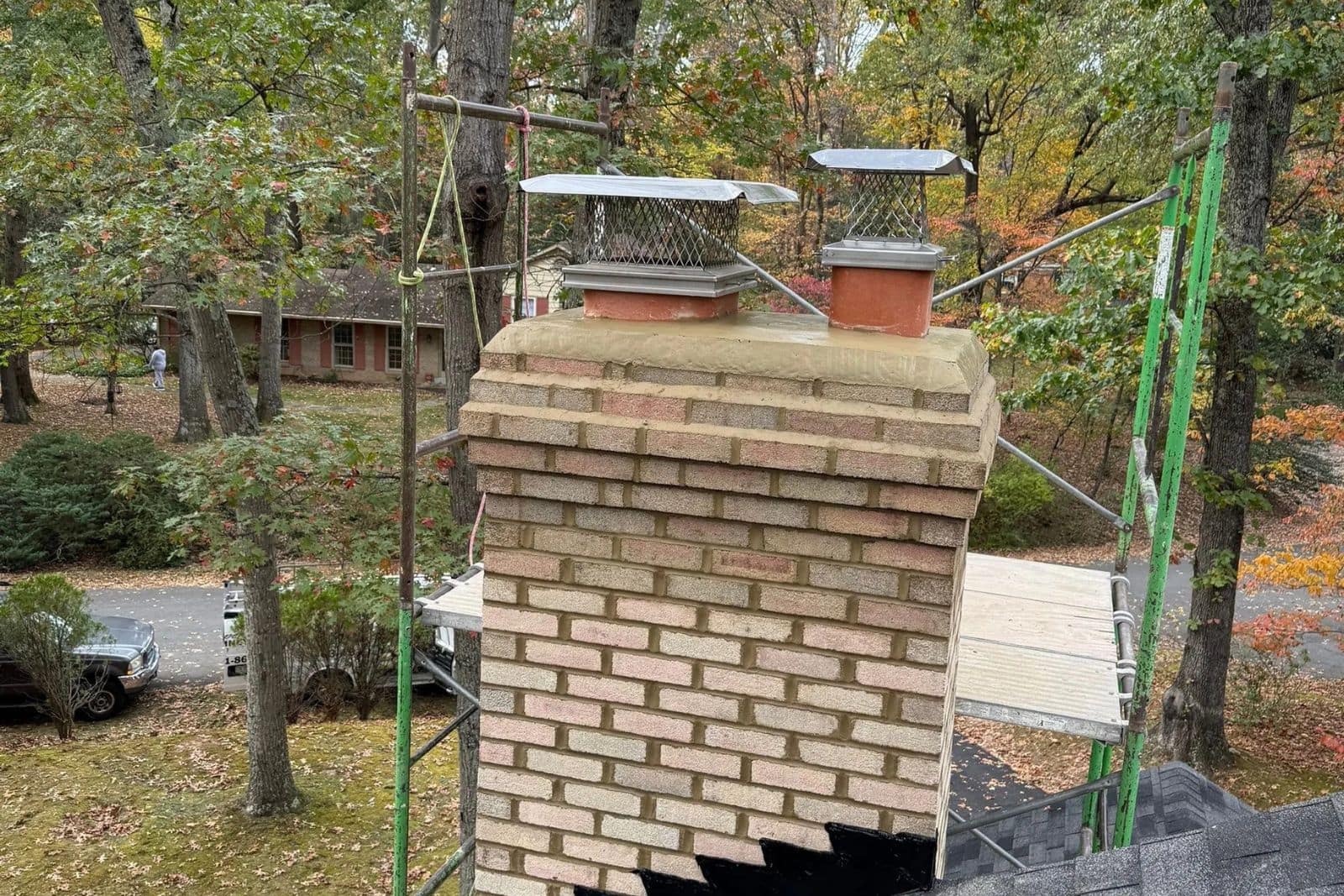 Scaffolding_against_chimney_during_repairs_in_reston_va_-__Sixpenny_Chimney_Sweeps Brick chimney with two metal caps and mesh guards, surrounded by autumn trees and scaffold.